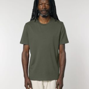 a man with dark skin, a beard and dreadlocks in a khaki green t-shirt and off-white trousers standing front on in front of a light grey background.