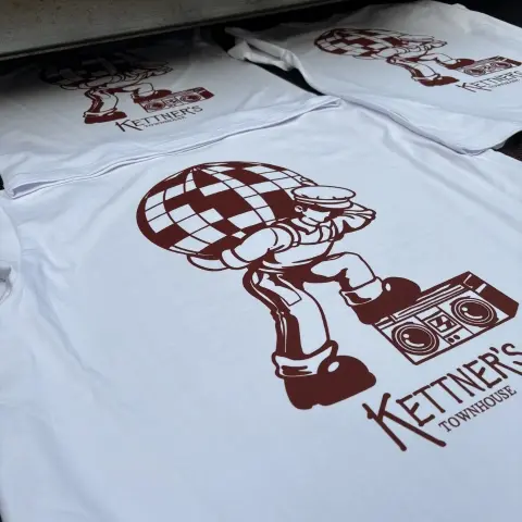Multiple white t-shirts with a screen printed disco style burgundy design, coming off of an industrial tunnel dryer.