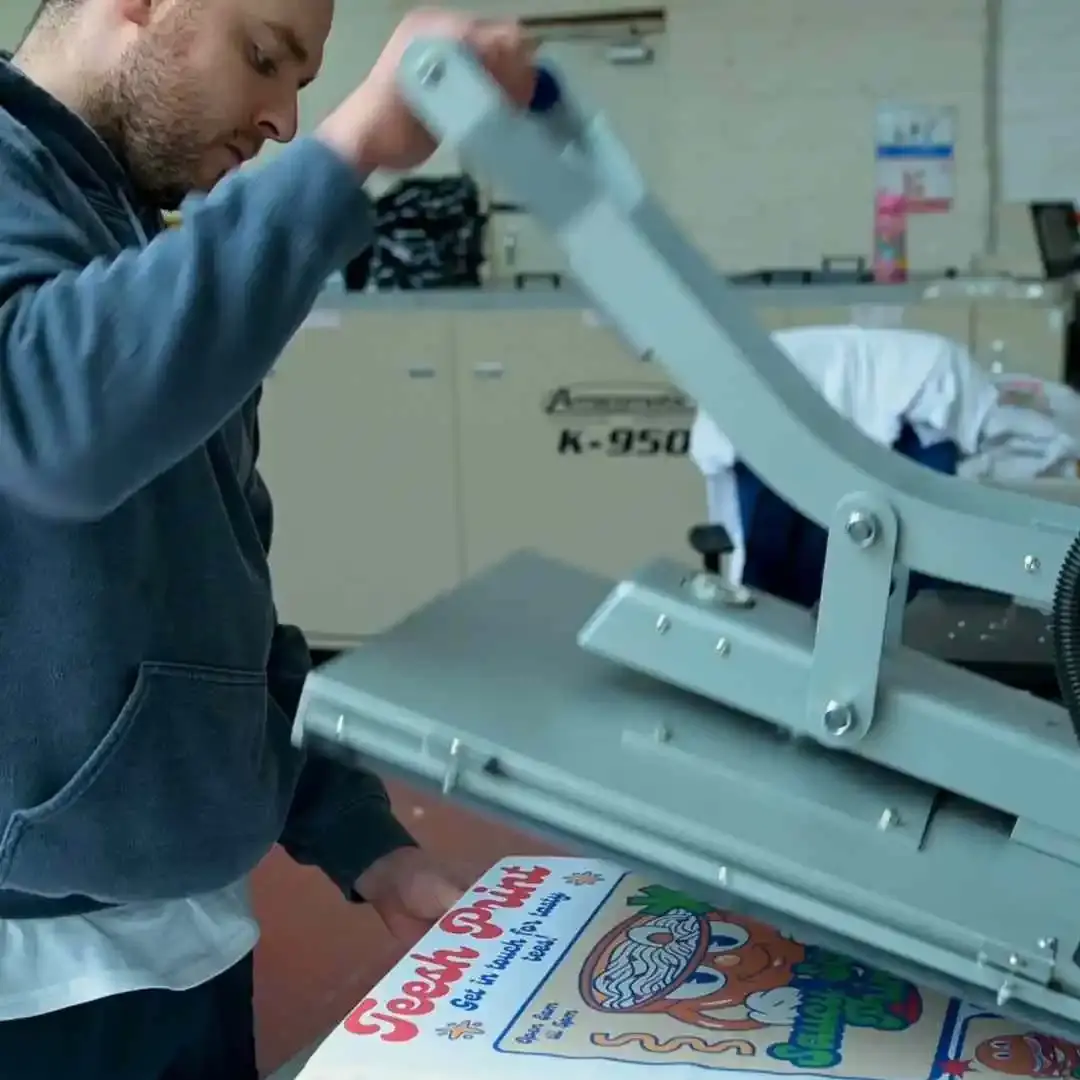 A garment printing professional using a heat press to apply a colorful transfer design onto a white T-shirt in a workshop setting.